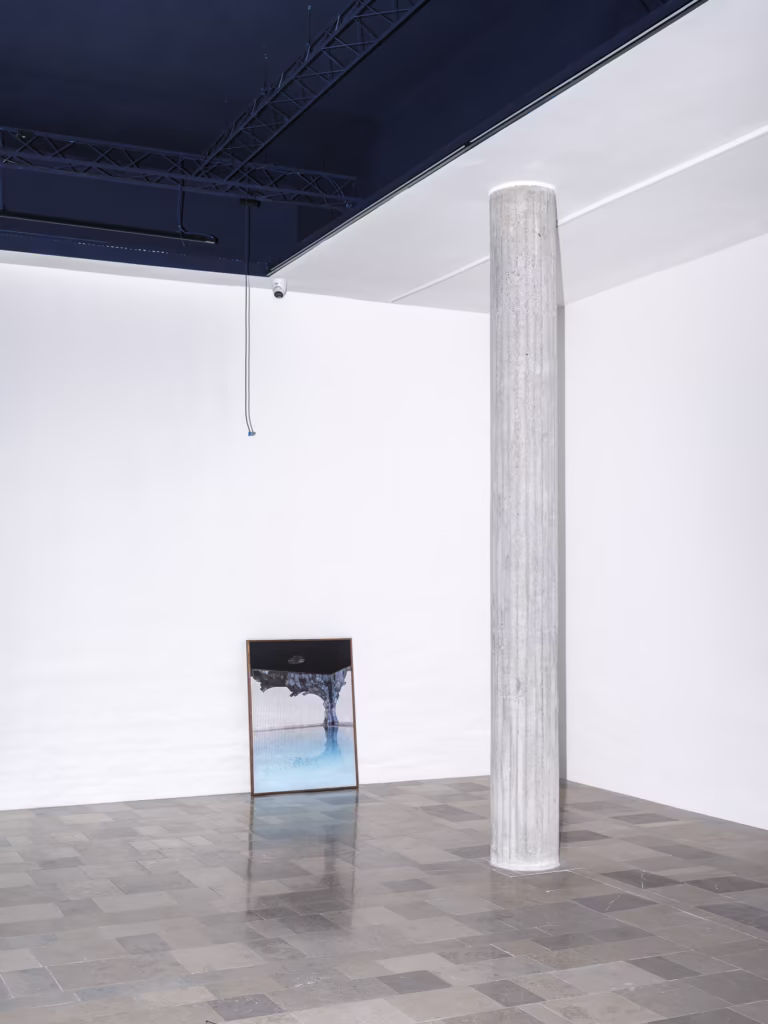 Betonowa kolumna stoi na wyłożonej kafelkami podłodze w pokoju o białych ścianach; oprawiona fotografia spoczywa pod ścianą pod odsłoniętym sufitem z kablami i zwisającym drutem.