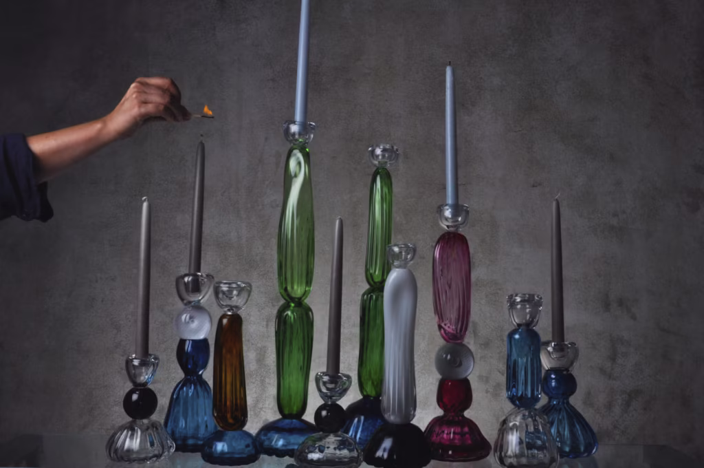 Ręka zapala niebieską świecę umieszczoną wśród różnokolorowych szklanych świeczników, z których każdy trzyma inną świecę, ustawionych na prostym szarym tle.