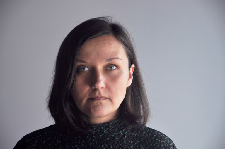 Kobieta z ciemnymi włosami do ramion i wzorzystym topem patrzy bezpośrednio w kamerę na prostym jasnoszarym tle.