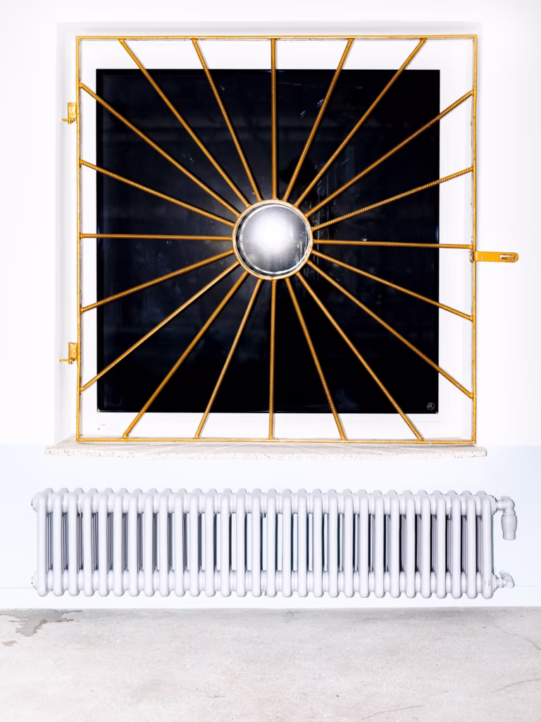 Kwadratowe okno z żółtymi metalowymi prętami we wzór słońca nad białym grzejnikiem, na tle białej ściany.