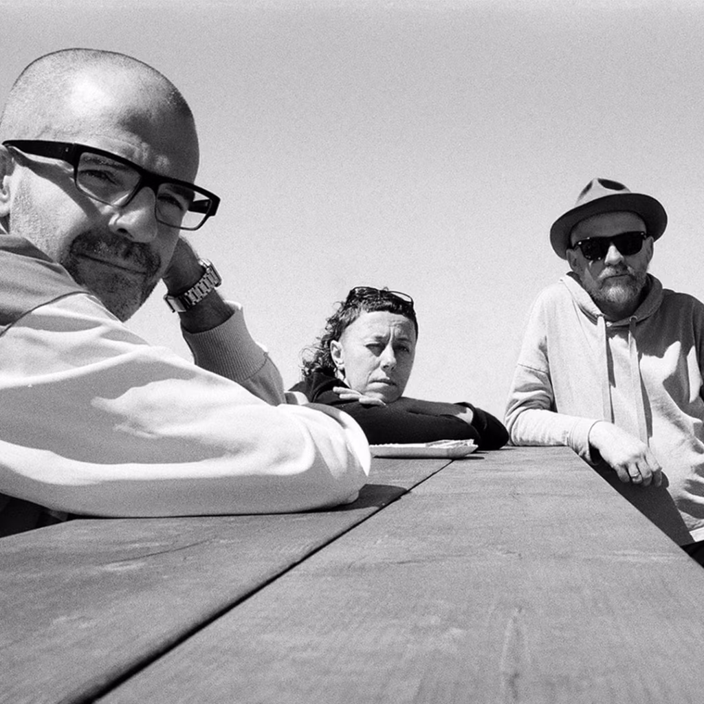 Trzy osoby siedzą przy drewnianym stole na świeżym powietrzu, patrząc w stronę aparatu. Zdjęcie jest czarno-białe i zostało zrobione pod niskim kątem.