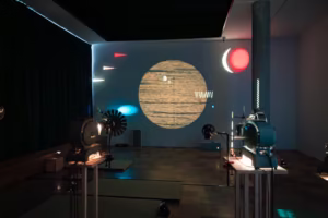 Ciemny pokój z zabytkowymi projektorami wyświetlającymi abstrakcyjne kształty i duże żółte koło na ścianie, obok rozproszonych kolorowych świateł i wzorów.