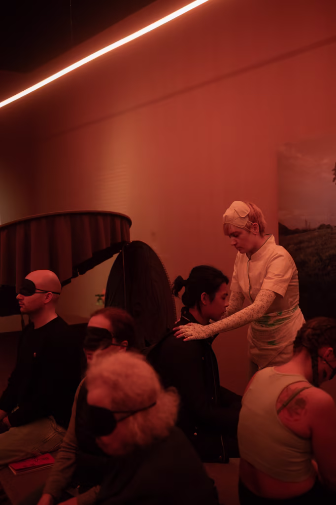 Osoba w białym stroju i rękawiczkach kładzie ręce na ramionach siedzącego uczestnika; inni w pobliżu siedzą z zawiązanymi oczami przy czerwonym oświetleniu.
