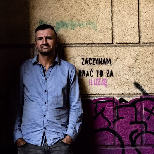 Mężczyzna w niebieskiej koszuli stoi pod ścianą z graffiti i polskim tekstem "Zaczynam brać to za iluzję".