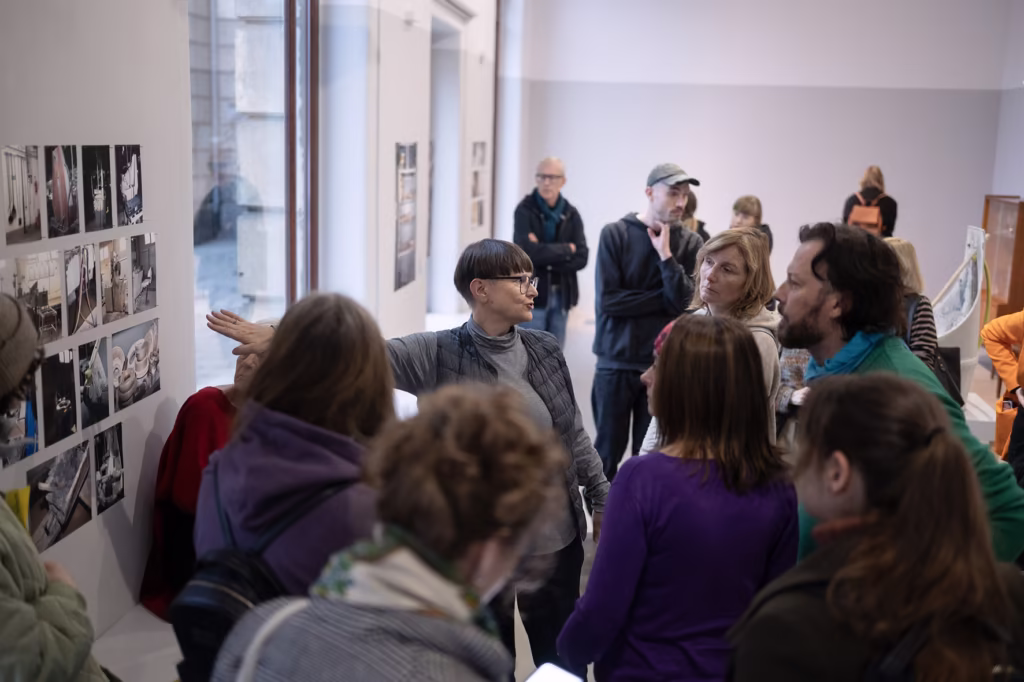 Kobieta gestykuluje w kierunku wystawy zdjęć na ścianie galerii, podczas gdy grupa ludzi uważnie słucha i obserwuje.