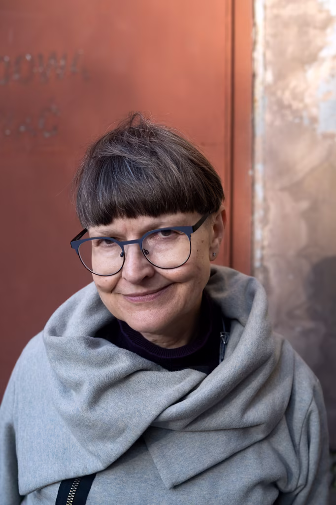 Kobieta o krótkich brązowych włosach i okularach, ubrana w szary szalik, stoi przed zwietrzałą czerwono-brązową ścianą, patrząc w kamerę z lekkim uśmiechem.