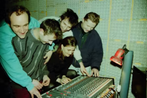 Pięć osób zebrało się wokół konsoli mikserskiej w dźwiękoszczelnym pomieszczeniu, regulując elementy sterujące i uśmiechając się.