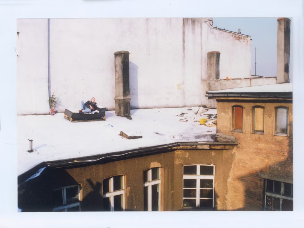Dwoje ludzi siedzi na kanapie na zaśnieżonym dachu obok kominów, a poniżej widać porozrzucane gruzy i stare fasady budynków.