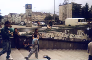 Kolorowe zdjęcie przedstawia schody do przejścia podziemnego, na których stoją i siedzą ludzie. W tle widać budynki i samochody. Na murze przyklejone są plakaty. W centrum kadru stoi mężczyzna z gitarą, przed nim na schodach leży biały kapelusz. Niektóre postaci uchwycone są w ruchu, schodzące po schodach.
