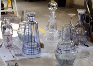 Kilka przezroczystych szklanych karafek i butelek z niebieskimi i czerwonymi wzorami siatki jest wyświetlanych na stole z papierami, na industrialnym tle.