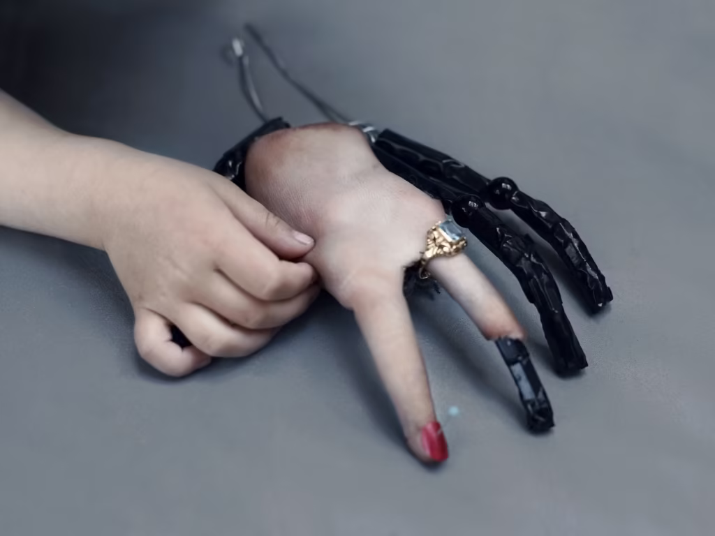 Ręka dziecka dotyka protezy dłoni z czarnymi mechanicznymi palcami, czerwonym lakierem do paznokci i złotym pierścieniem, spoczywającej na szarej powierzchni.