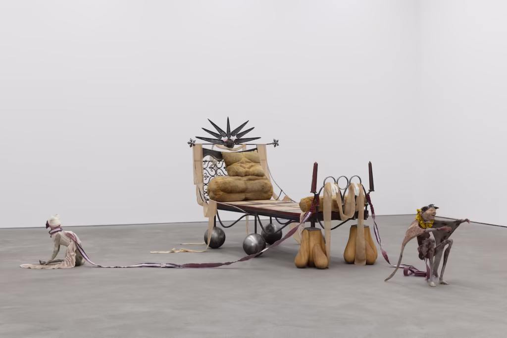 Rzeźbiarska instalacja przedstawiająca dekoracyjny drewniany tron na kółkach, z abstrakcyjnymi postaciami przypominającymi ludzi, ciągniętymi za pomocą długich pasów tkaniny, wyświetlana w otoczeniu galerii.