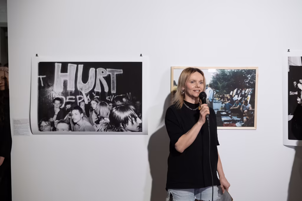 Kobieta mówi do mikrofonu przed ścianą prezentującą czarno-białe i kolorowe fotografie na wystawie sztuki.