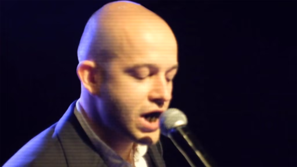 Osoba z ogoloną głową śpiewa lub mówi do mikrofonu na scenie, ubrana w ciemną kurtkę, na ciemnym tle.