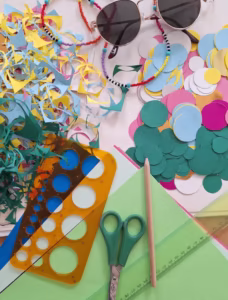 Różne papierowe wycinanki, ołówek, nożyczki, linijka, szablon i okulary przeciwsłoneczne z koralikami ułożone na powierzchni, sugerujące projekt artystyczny i rzemieślniczy.