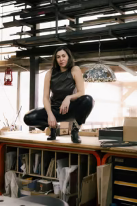 Kobieta w czarnym skórzanym ubraniu siedzi na drewnianym stole roboczym w warsztacie, otoczona półkami, narzędziami i różnymi materiałami.