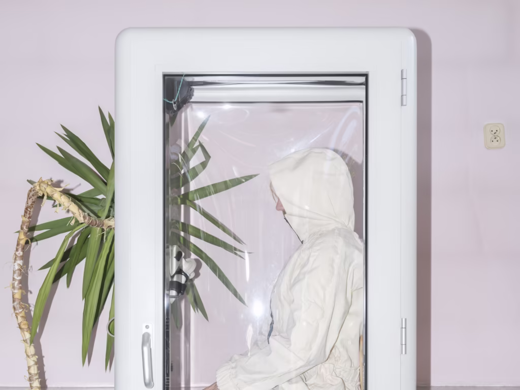 Osoba w białym kombinezonie z kapturem siedzi wewnątrz przezroczystej obudowy przypominającej budkę telefoniczną obok rośliny doniczkowej na tle bladoróżowej ściany.