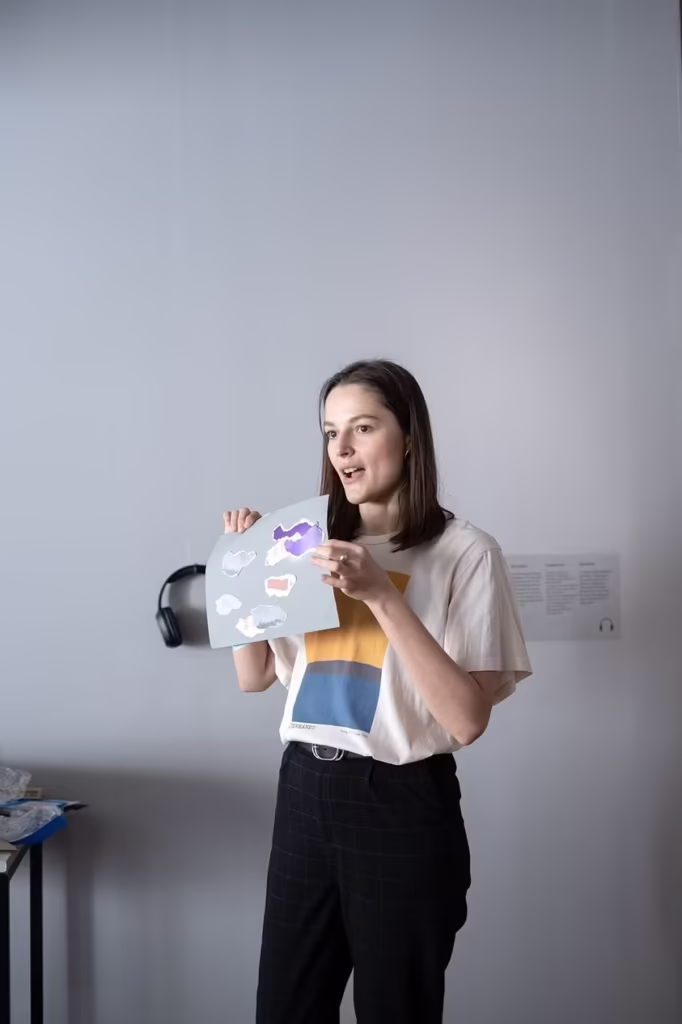 Kobieta stoi w pomieszczeniu, trzymając arkusz z kolorowymi kształtami, zdając się mówić lub prezentować przed jasnoszarą ścianą.