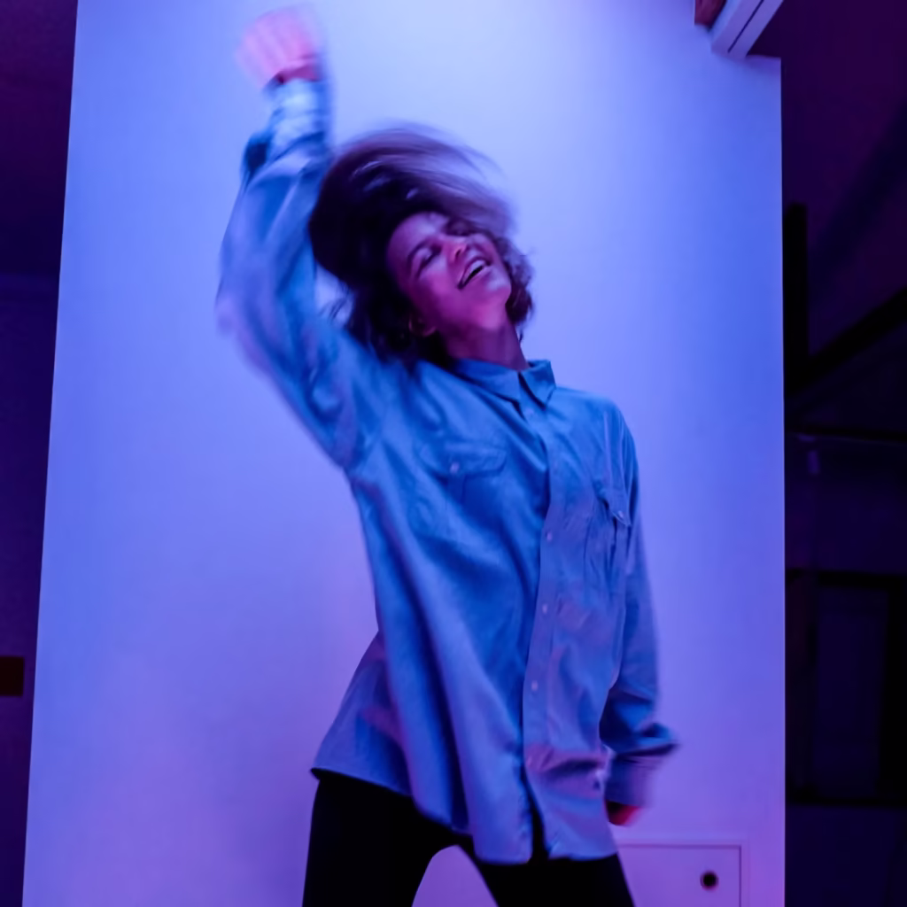 Osoba w za dużej niebieskiej koszuli tańczy energicznie w fioletowo-niebieskim oświetleniu, z głową odchyloną do tyłu i włosami kołyszącymi się w ruchu.