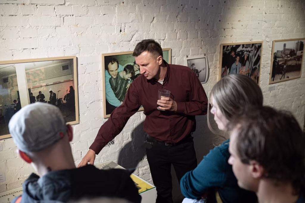 Mężczyzna z drinkiem gestykuluje w kierunku wyświetlacza, przemawiając do małej grupy przed oprawionymi fotografiami na ścianie z białej cegły.