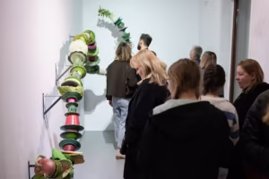Ludzie oglądają współczesną instalację artystyczną przedstawiającą ceramiczne lub plastikowe naczynia i miski przymocowane do ściany w białej przestrzeni galerii.