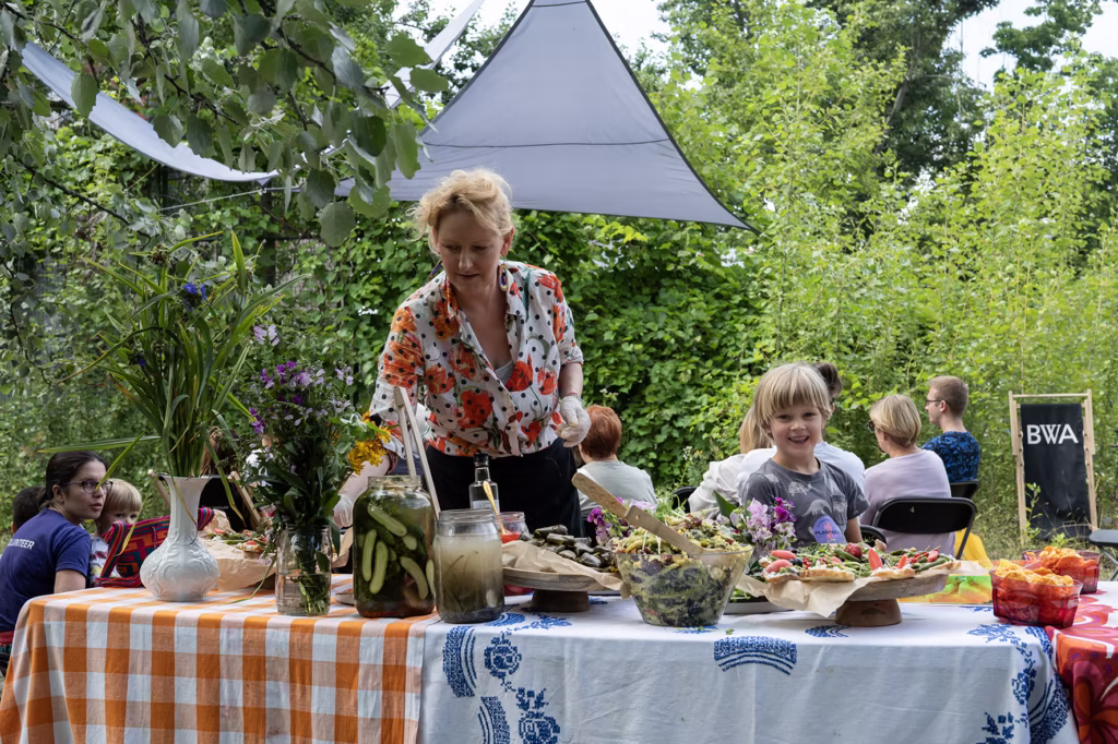 Kobieta i dziecko stoją obok stołu z różnymi potrawami i napojami podczas spotkania na świeżym powietrzu, z innymi osobami siedzącymi w tle wśród zieleni.