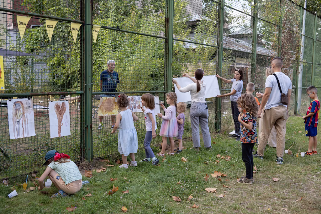Dzieci i dorośli stoją przy drucianym ogrodzeniu na zewnątrz, wieszając i oglądając prace plastyczne dzieci przymocowane do ogrodzenia za pomocą spinaczy do bielizny. Niektóre dzieci malują lub rysują na trawie.