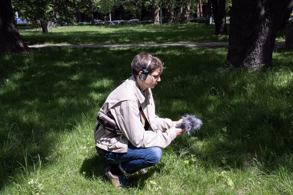 Osoba w słuchawkach przykuca na trawie w parku, trzymając przenośny rejestrator dźwięku z przednią szybą.