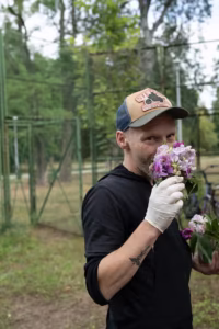 Mężczyzna w czapce i białej rękawiczce trzyma i wącha bukiet fioletowych kwiatów na zewnątrz w pobliżu zielonego ogrodzenia i drzew.