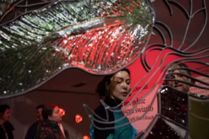 Kobieta stoi w pobliżu dekoracyjnej szklanej rzeźby z czerwonym i zielonym oświetleniem; w tle widoczne są inne osoby i tekst na ścianie.