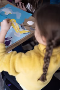 Dziecko z zaplecionymi włosami w żółtym swetrze tworzy kolaż z podartego kolorowego papieru przy stole.