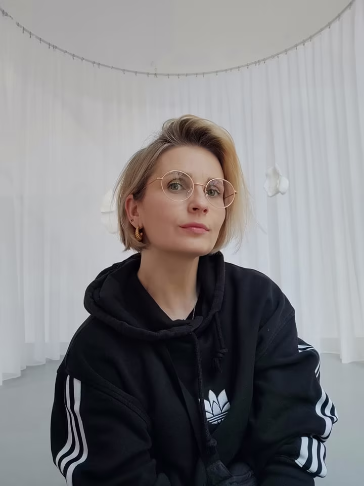 Osoba o krótkich blond włosach i okularach w czarnej bluzie z kapturem siedzi przed białymi zasłonami w dobrze oświetlonym pokoju.