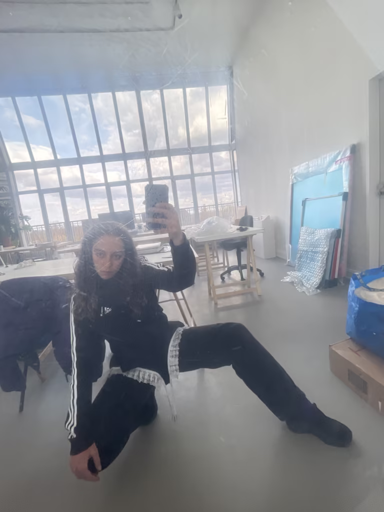 Osoba w czarnej odzieży sportowej robi sobie selfie w lustrze w przestronnym, dobrze oświetlonym pokoju z dużymi oknami, materiałami artystycznymi i materiałami do pakowania w tle.