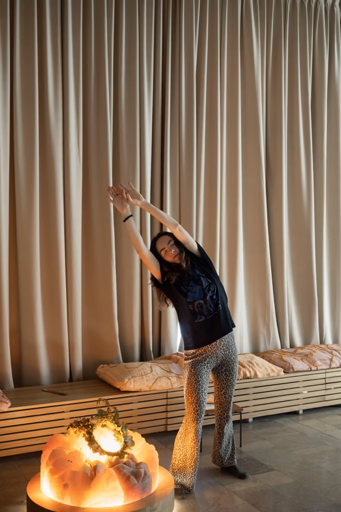W pokoju z beżowymi zasłonami, ławką do siedzenia i świecącą lampą na stole osoba rozciąga się, wyciągając ręce nad głowę i na boki.