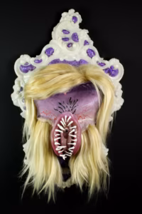 Ozdobna ramka przedstawia surrealistyczną maskę z blond włosami, fioletową twarzą, ostrymi białymi zębami i wyraźnymi ustami pośrodku, na czarnym tle.