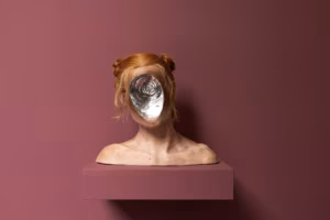 Rzeźba przedstawiająca głowę osoby o czerwonawych włosach, upiętych w dwa koki, ma w miejscu twarzy lustrzane, spiralne wgłębienie; stoi na dopasowanym cokole przed fioletowo-różową ścianą.