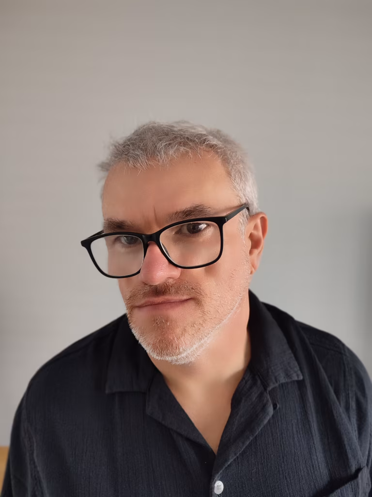 Mężczyzna o krótkich siwych włosach, z lekkim zarostem i w czarnych okularach ma na sobie czarną koszulę z kołnierzykiem i patrzy w obiektyw przed jednolitym tłem.