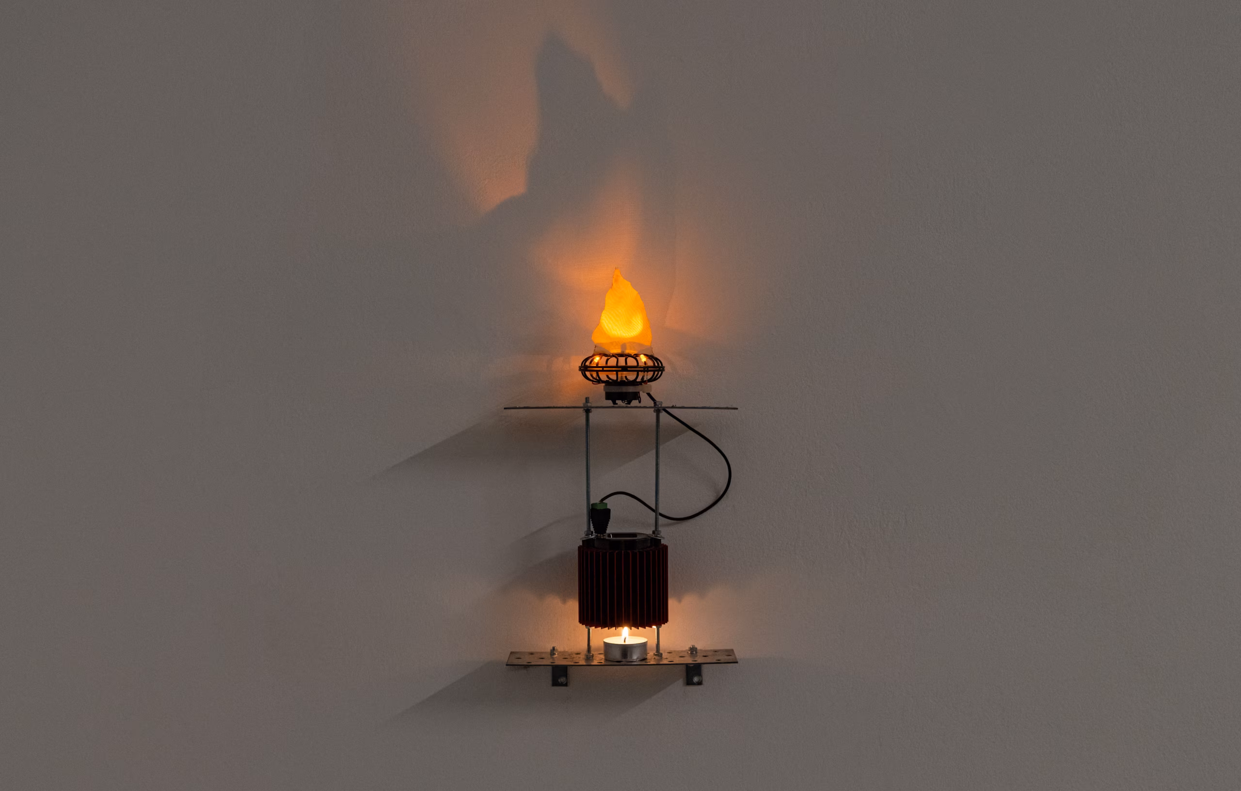 Urządzenie montowane na ścianie, w którego dolnej części umieszczono zapaloną świeczkę typu tealight, a w górnej – świecącą żarówkę w kształcie płomienia.